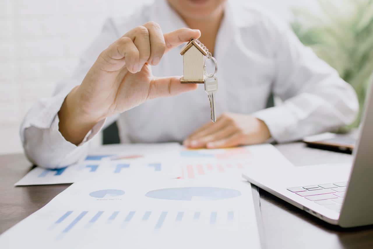 Woman in White Long Sleeve Shirt Holding House Shaped Key Chain Sitting On Desk With Papers And Laptop